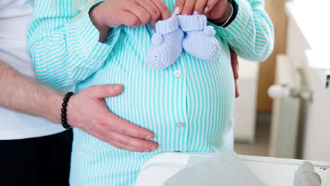 future mom holding blue knitted socks stepping them by the big belly. male hands stroking the abdomen of wife. close up.