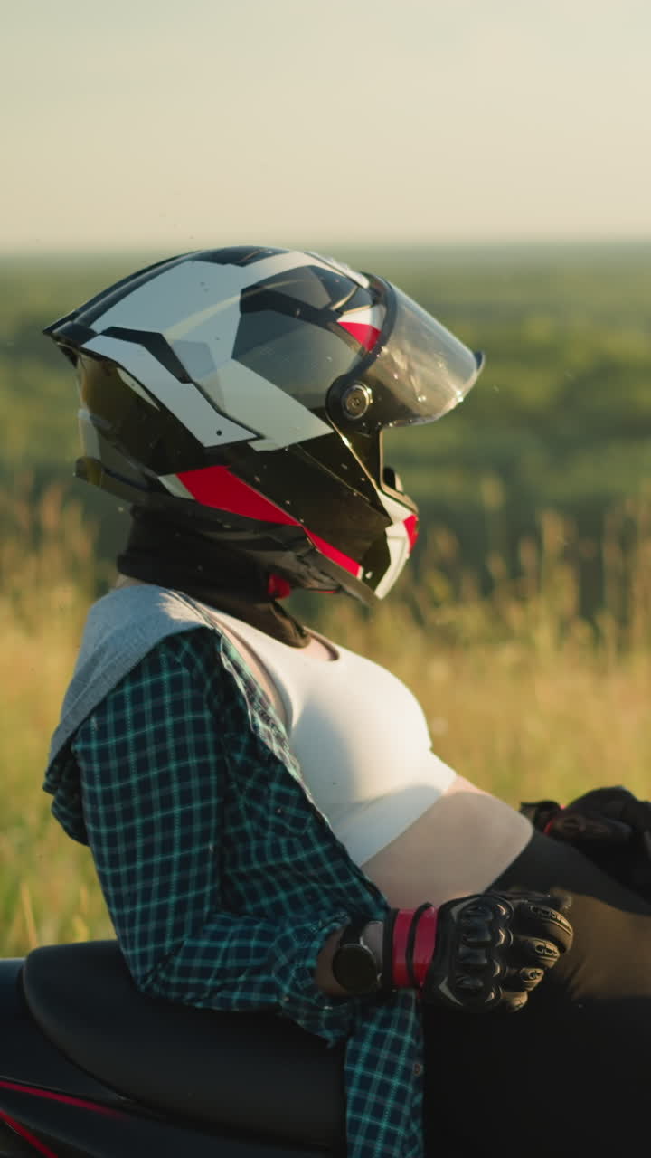 A young woman wearing a helmet rests on a red power bike in a field, adjusting her position slightly forward, her white crop top is visible, while the wind gently moves the surrounding grass