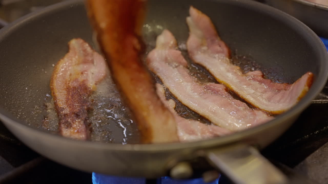 Listarelle di bacon che sfrigolano in una padella. Scena ravvicinata di preparazione con consistenza croccante che si dora, grasso bollente e vapore che si alza su un fornello: preparazione classica di cibo per la colazione in cucina.