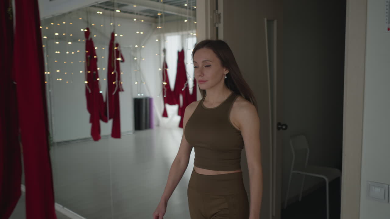 Yoga specialist walking into practice room with poised posture and flowing motion under soft natural light, framed by hanging red hammocks and purple curtains creating serene fitness atmosphere