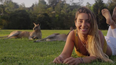 girl smiling at camera while comfortably lying on the green grass together with eastern grey kangaroos - gold coast, qld, australia