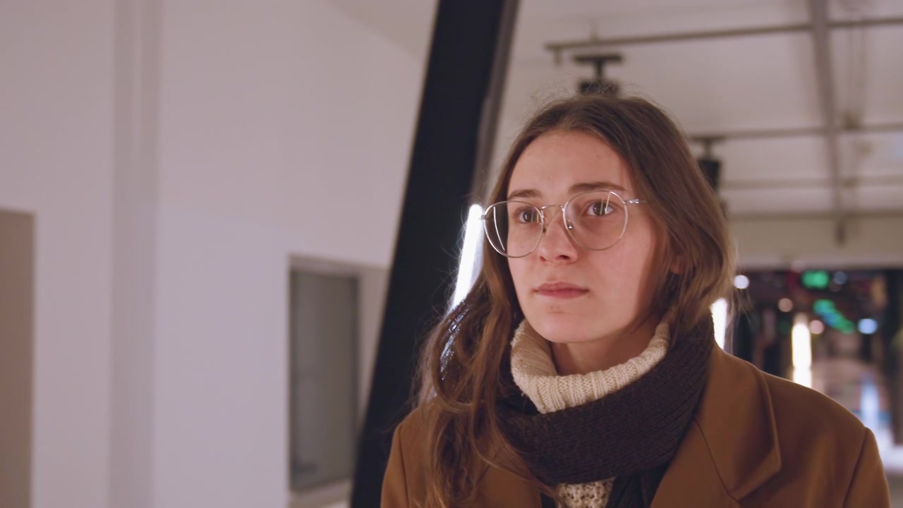 White woman standing inside lit corridor, young white artist in brown coat and glasses pausing to study architectural lines, back and profile shots, warm LED accents and angled beams, quiet resolve