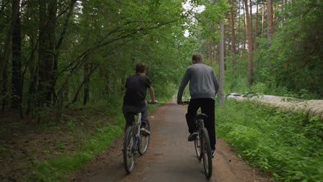 two friends riding bicycles along a paved path surrounded by lush green trees, one is standing while cycling, enjoying the fresh air and nature, with electric pole by the side