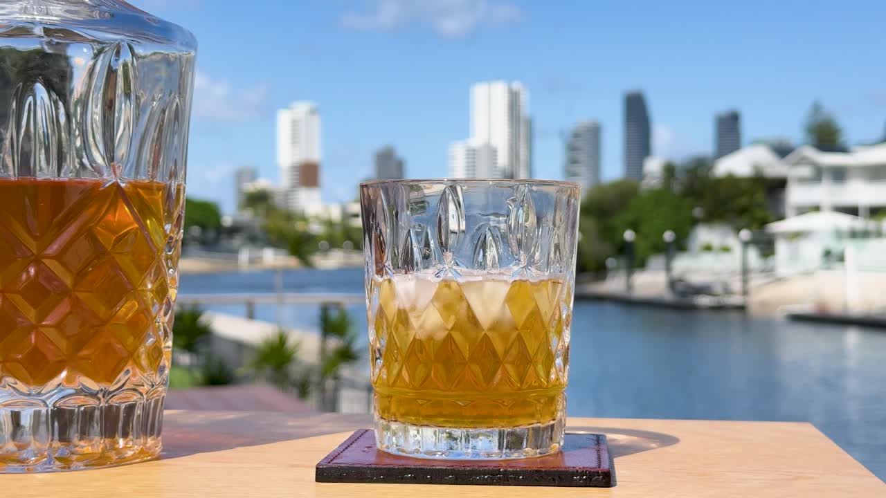 A hand lifts a crystal whiskey glass with ice from a wooden table, set against a bright, sunny waterfront city skyline. Static camera, natural daylight