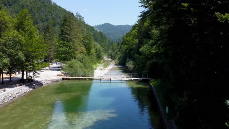 Aerial-flyover-clear-lake,-bridge-and-natural-river-Surrounded-by-green-Forest-trees-in-Global-Geopark-Idrija-during-sunny-day-,-Slovenia