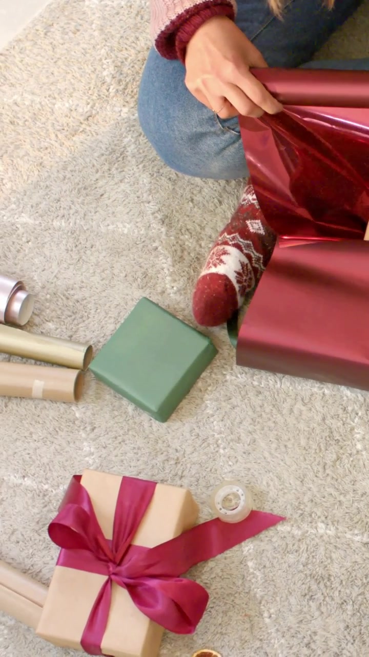 Video vertical: Desenrollando un rollo burdeos, mujer en la alfombra envolviendo un regalo en casa con suéter y jeans