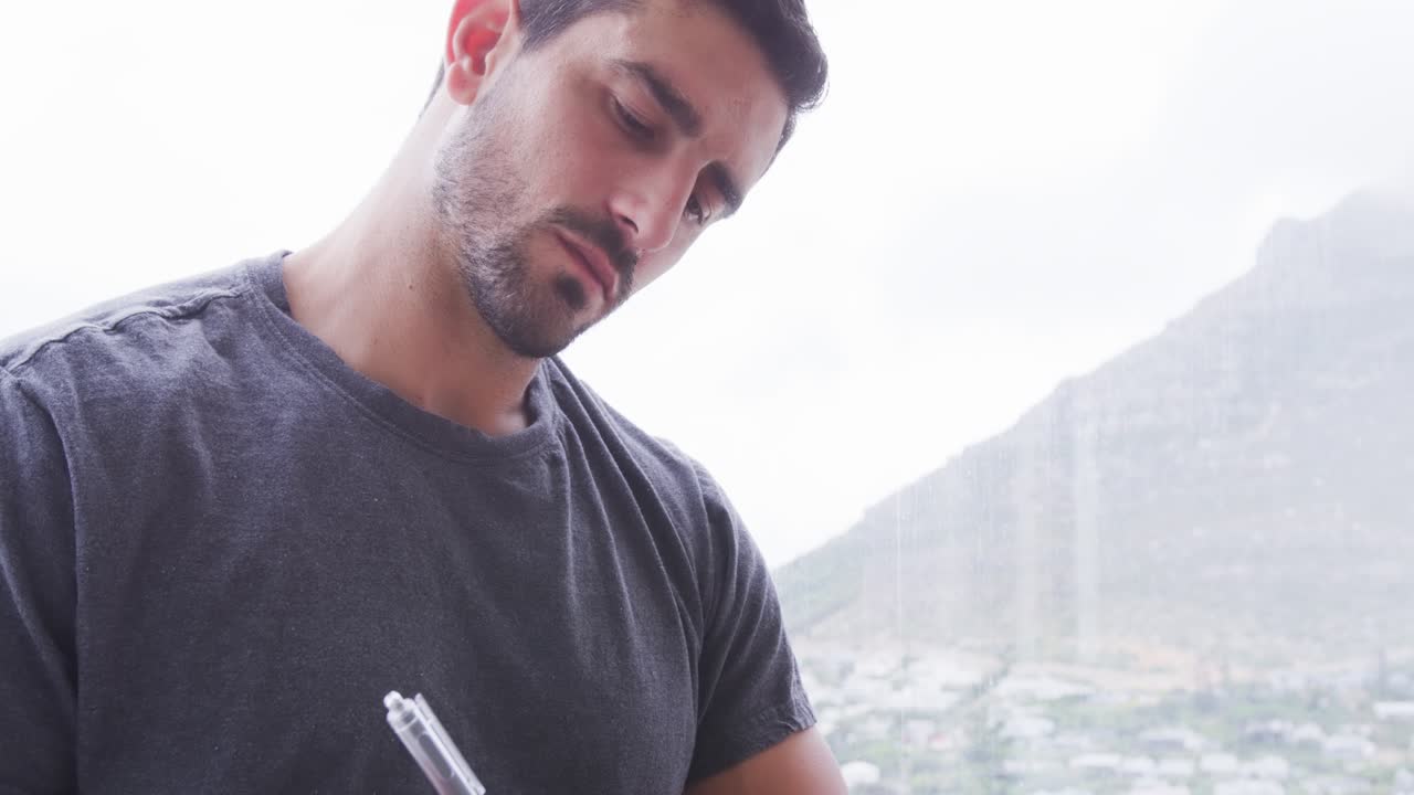 Homem encontrando uma página no caderno, batucando a caneta e escrevendo um diário ao lado da janela com vista para a montanha e uma pulseira.