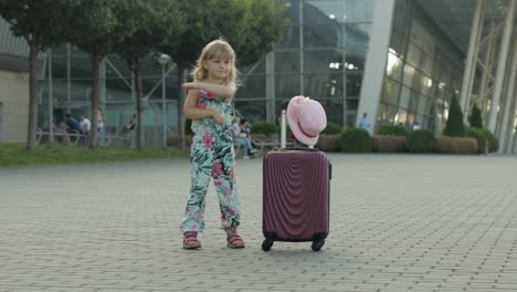 child girl tourist with suitcase luggage bag near airport. kid dances, rejoices, celebrates