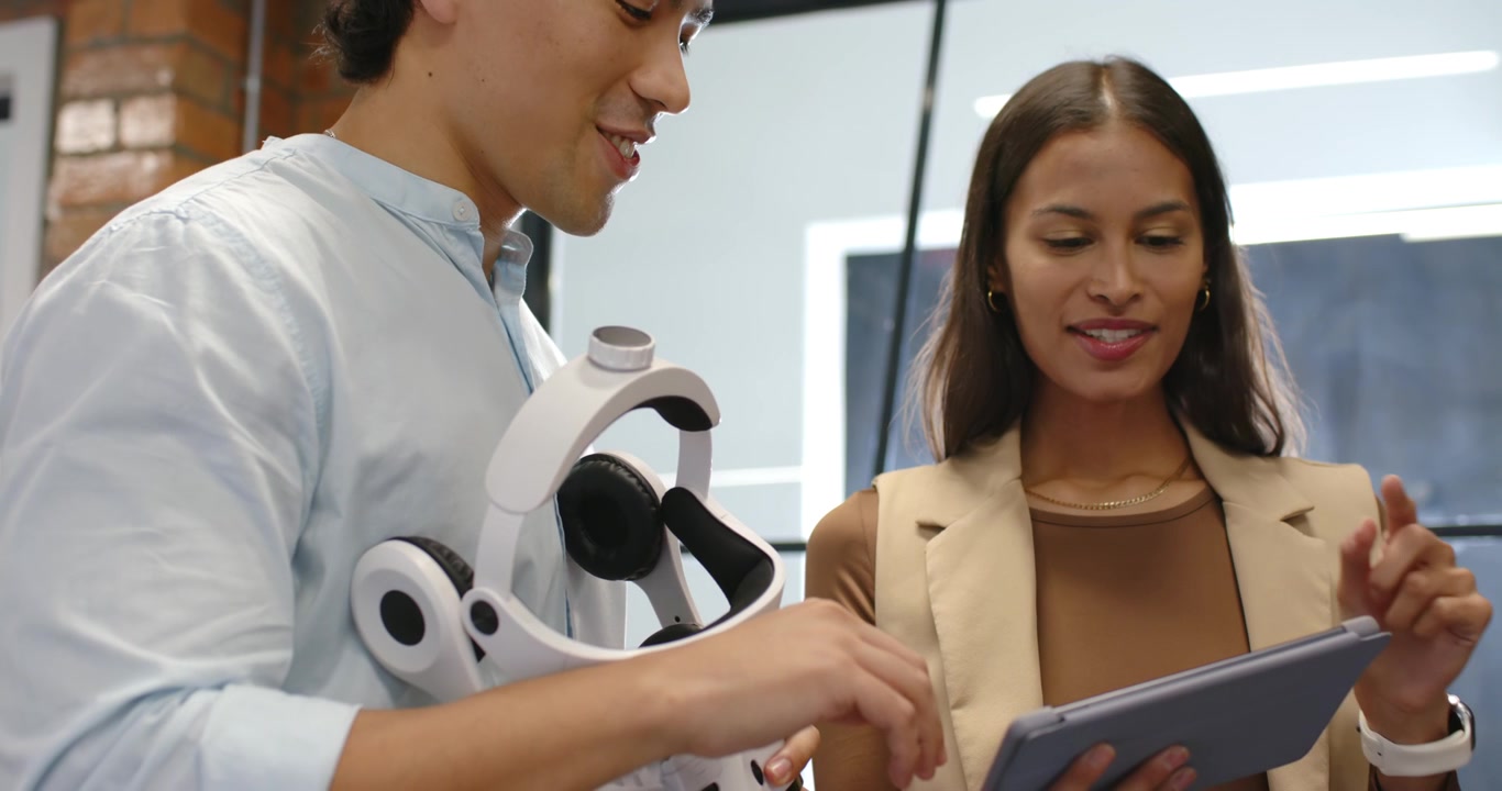 Diverse coworkers in office, man speaking explaining headset, woman tapping tablet navigating