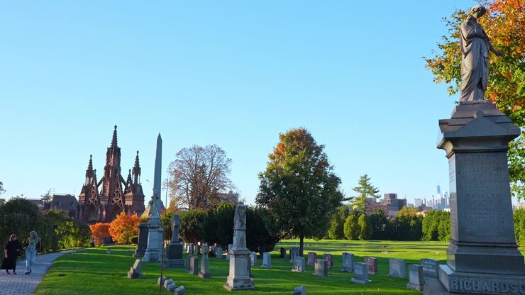 New York, USA, 8 ottobre 2026: monumenti cimiteriali fiancheggiano un prato con cancelli gotici in lontananza. Una statua si erge sulla destra mentre i visitatori seguono un sentiero accanto al fogliame arancione sotto un cielo blu