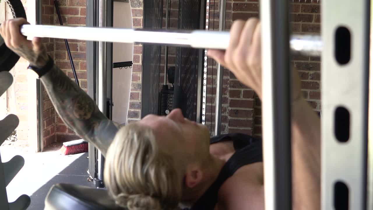 Tattooed muscly man in home gym working out bench press barbell