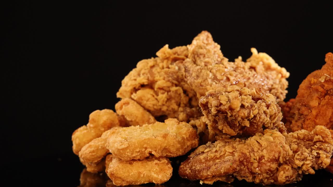 Assorted fried chicken wings, drumsticks, and nuggets rotate on a reflective black surface under warm, studio lighting, highlighting crunchy textures and golden color