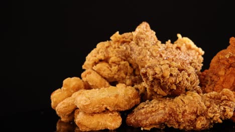 assorted fried chicken wings, drumsticks, and nuggets rotate on a reflective black surface under warm, studio lighting, highlighting crunchy textures and golden color