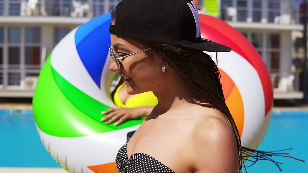 dos chicas atractivas con gafas de sol caminando con tubos inflables junto a la piscina y sonriendo. mujeres bonitas teniendo una fiesta en la piscina. disparo en cámara lenta