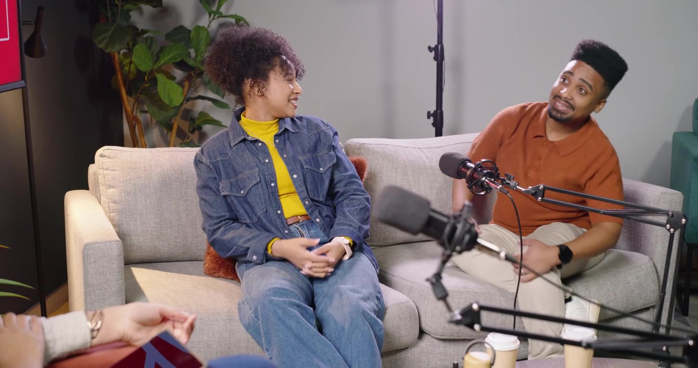 Conduttori afroamericani, donna in jeans e uomo con polo color ruggine, che si girano gesticolando agli applausi in studio con microfono in registrazione