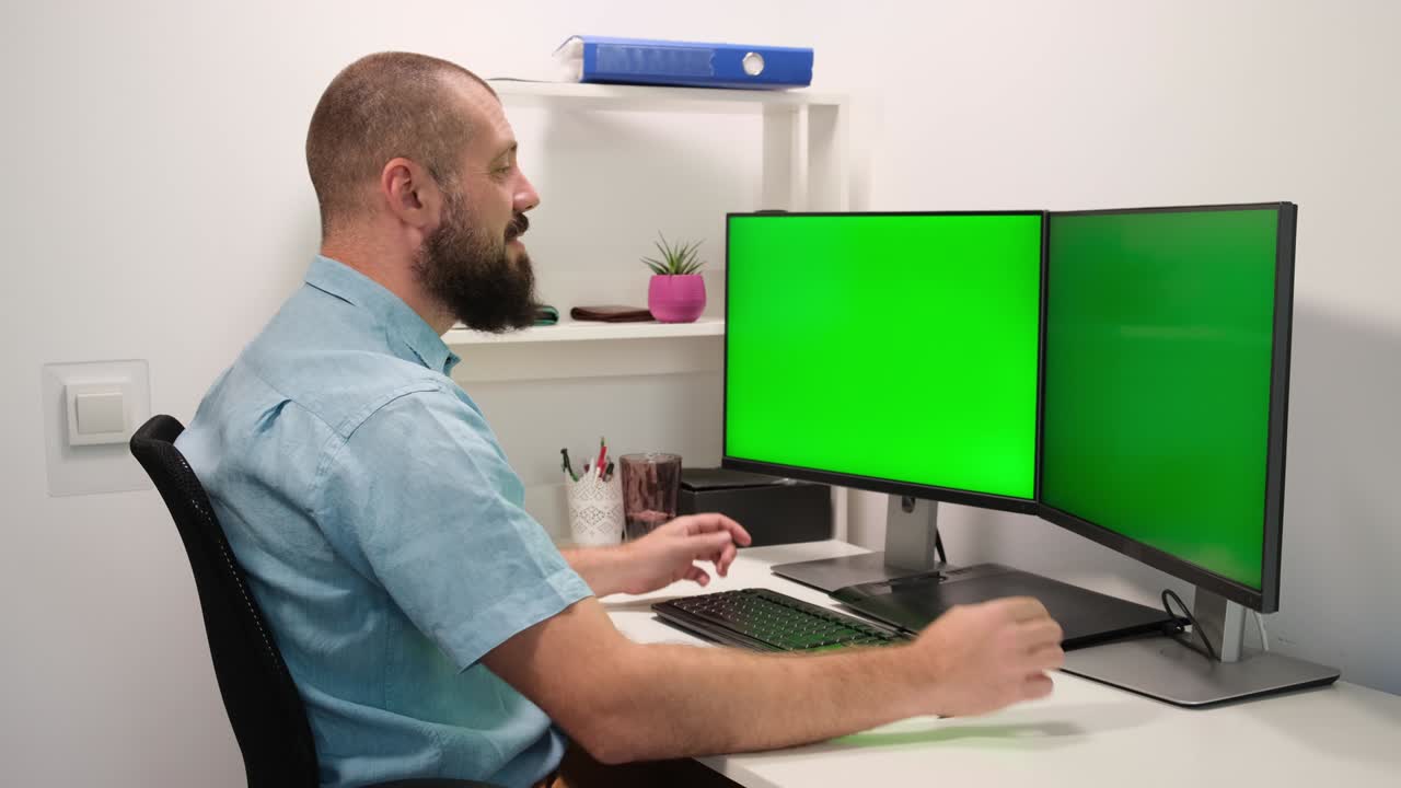 el oficial masculino trabaja en una computadora con dos maquetas de pantalla verde en casa. los empleados se sientan frente a las pantallas.