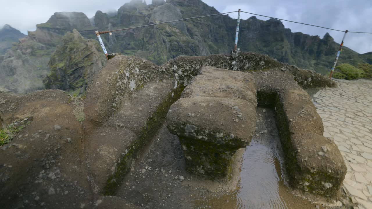 Weathered stone basins at Pico do Arieiro viewpoint, Madeira. Mist drifts over dramatic mountain ridges as the camera glides past carved rocks and rain pools, revealing the rugged summit