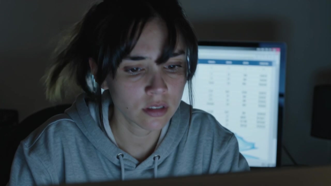 Leaning woman in gray hoodie responding to spreadsheet on rear monitor, analyzing data with laptop