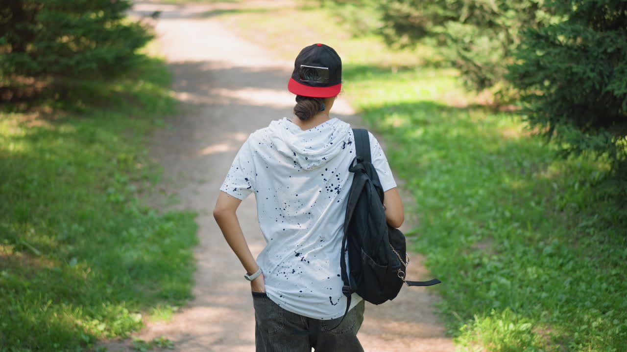 persona joven caminando por un sendero en un parque, con una mochila al hombro, gorra de visera roja y sudadera con capucha de lunares, luz solar moteada entre los árboles, ritmo relajado y estado de ánimo introspectivo, vegetación veraniega y suave