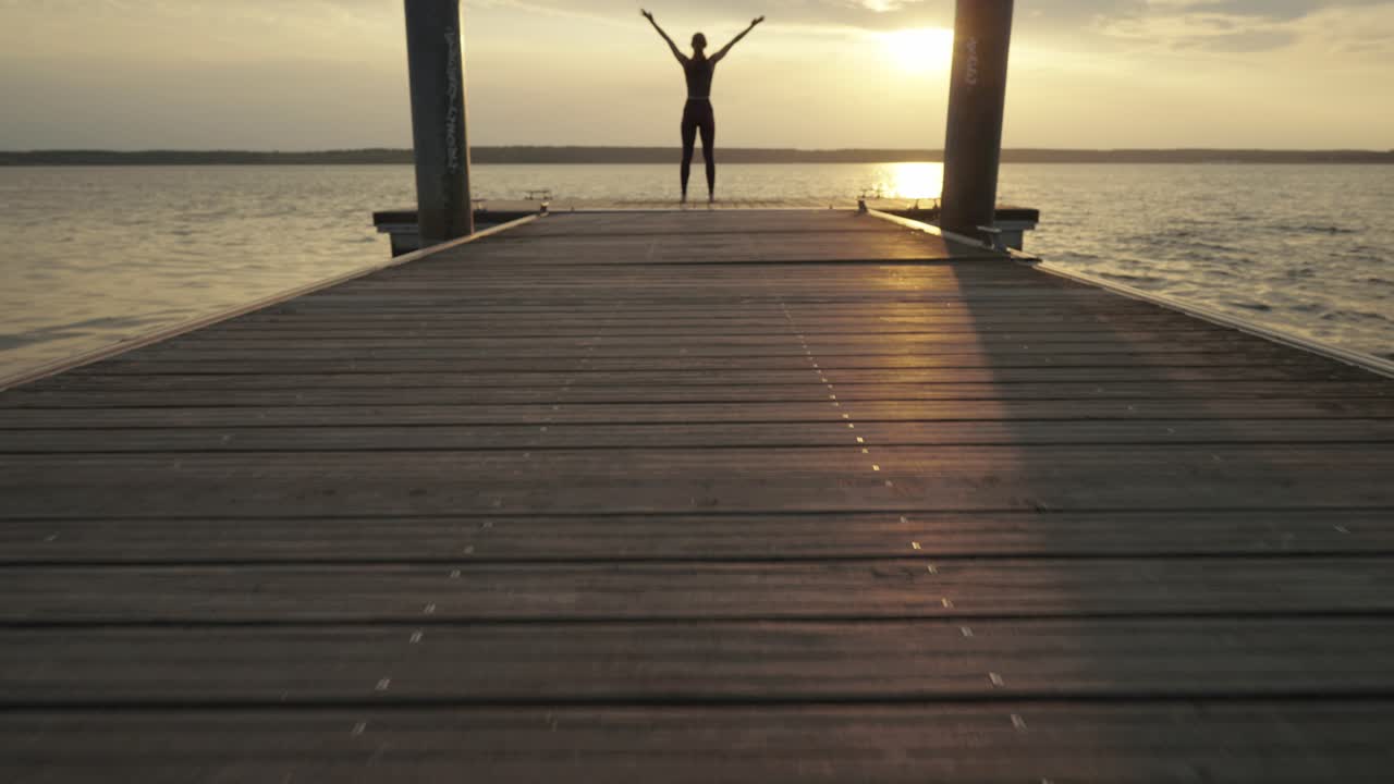 mujer joven haciendo yoga mientras está de pie en el embarcadero al atardecer, cámara lenta