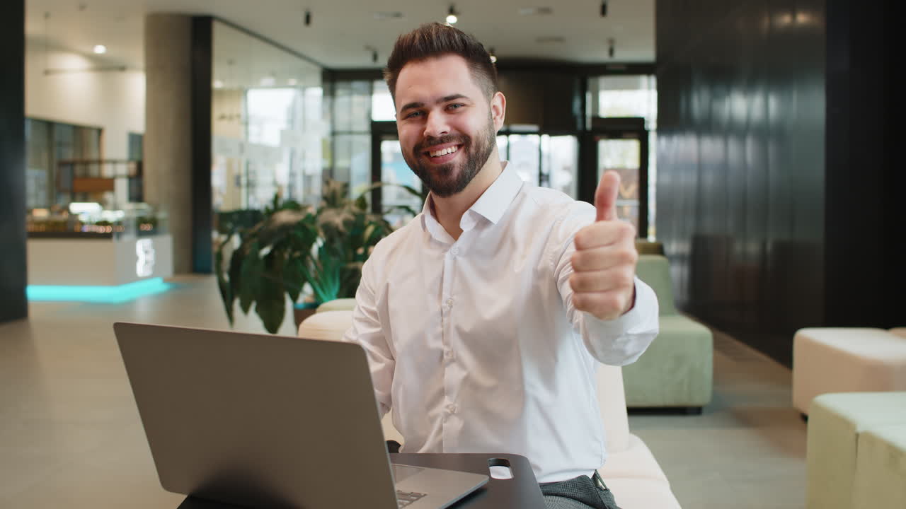 Happy young businessman working on laptop looking at camera showing thumbs up in modern office lobby