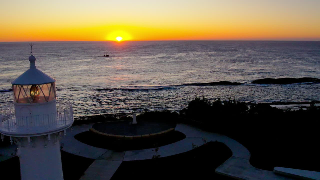 Drone flies toward the horizon over Warden Head Lighthouse at sunrise near Ulladulla, NSW. A fishing boat cruises through the calm ocean, adding life to this peaceful coastal morning scene.