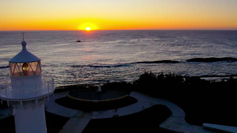 drone flies toward the horizon over warden head lighthouse at sunrise near ulladulla, nsw. a fishing boat cruises through the calm ocean, adding life to this peaceful coastal morning scene.