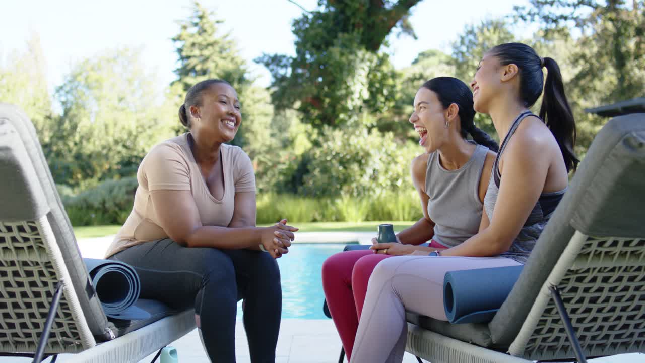 Diverse female friends in sportswear, left woman speaking after yoga poolside, passing cup laughing