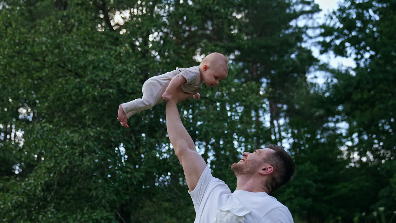 Muscular Caucasian man holding his newborn above head with one hand. Smiling happy dad entertaining his son. Nature backdrop.