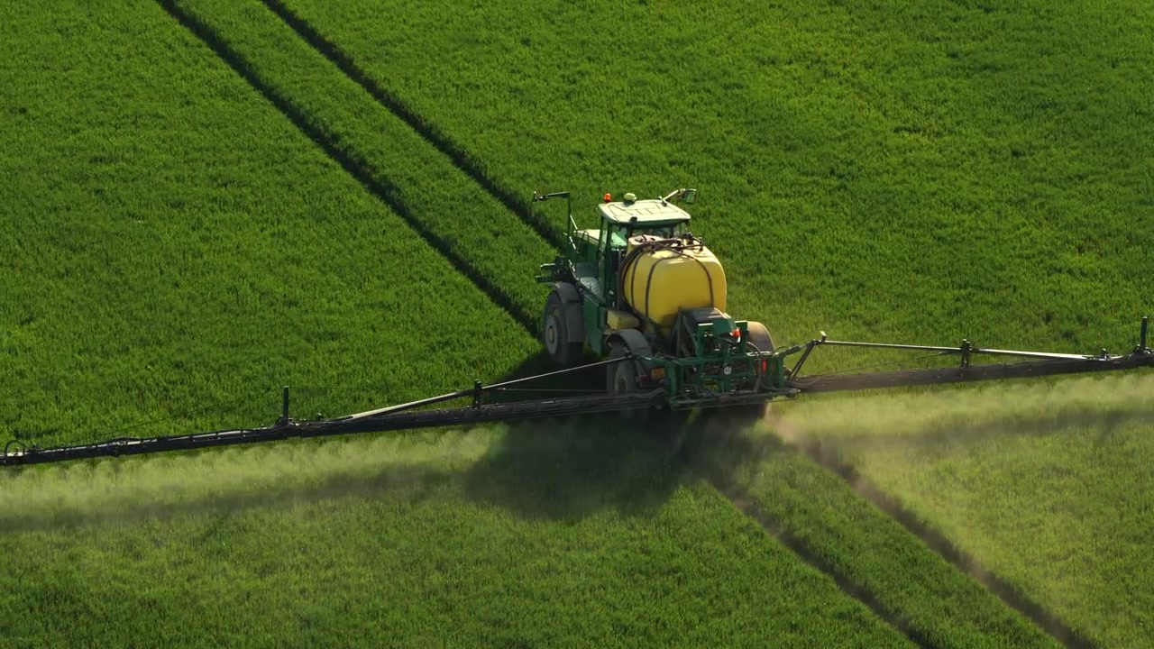 Vista aerea con drone di un trattore irroratore su un campo verde in collina, con colline boscose ondulate e valli agricole sullo sfondo, Moravia, Repubblica Ceca.
