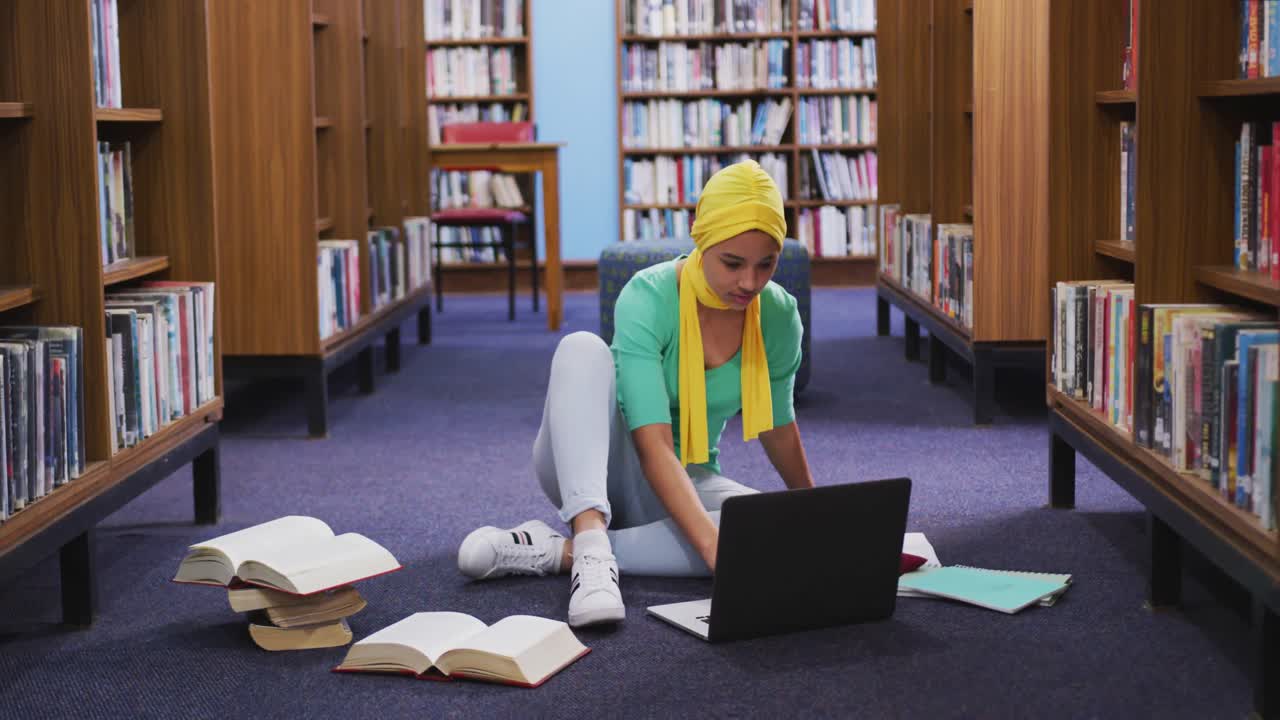 una estudiante asiática con un hijab amarillo estudiando en una biblioteca y usando una computadora portátil