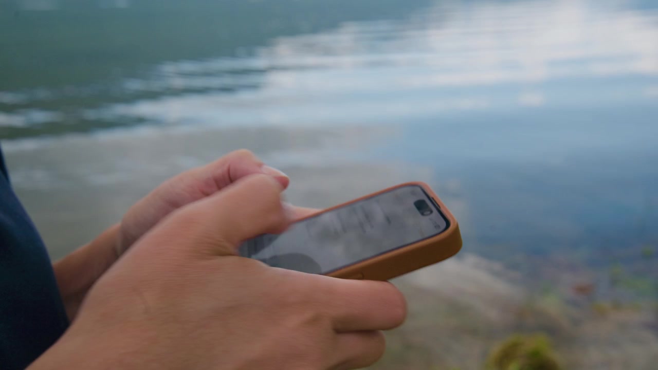 Mano tiene il telefono vicino all'acqua. Persona controlla messaggi o scatta foto. Momento calmo e concentrato in riva al lago. Ottimo per contenuti di viaggio, tecnologia o natura. Adatto per annunci, blog o social media