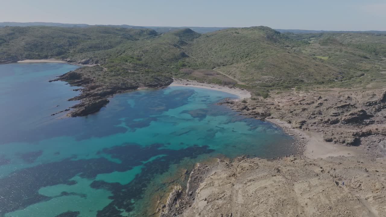Volo di drone aereo sopra l'incontaminata spiaggia di Cala Sa Torreta sull'isola di Minorca, costa spagnola, Paesaggio naturale, mare blu, colline verdi intorno alla costa sabbiosa e rocciosa