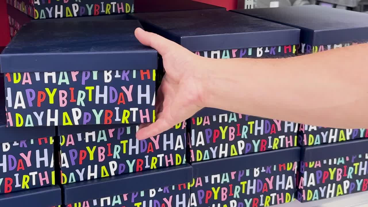 A hand reaches for a colorful 'Happy Birthday' gift box on a store shelf.