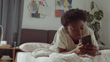 Mujer-Negra-Alegre-Acostada-En-La-Cama-Y-Usando-Un-Teléfono-Inteligente