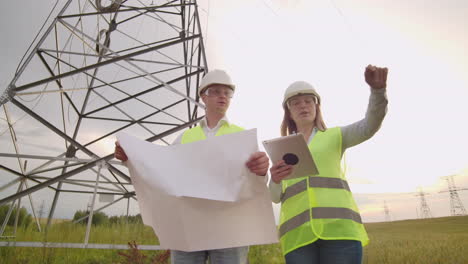 an electrician male and female in the fields near the power transmission line. he is an electrician who manages the process of erecting power lines. the mechanic in a helmet and manager with tablet