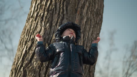 a young boy leaning his back against a large tree trunk, looking upward, with mouth open and his tongue touches his upper teeth with both arms up, he is dressed in a black jacket