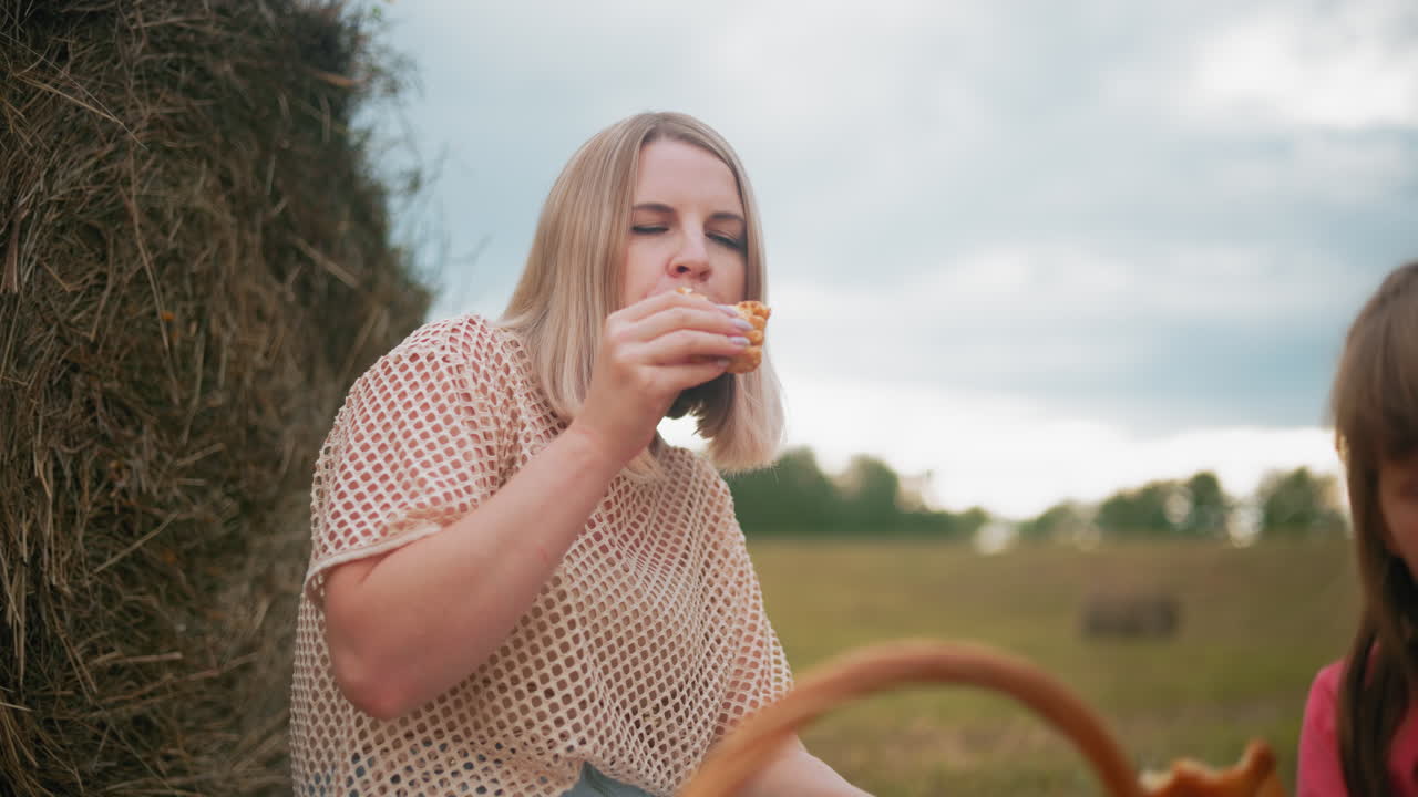 어린 어른은 페이스트리를 즐기고, 아이들은 근처에 앉아서 무언가에 집중합니다. 광활한 농장 배경 속에서 피크닉 바구니가 평화로운 야외 가족 순간을 만니다.