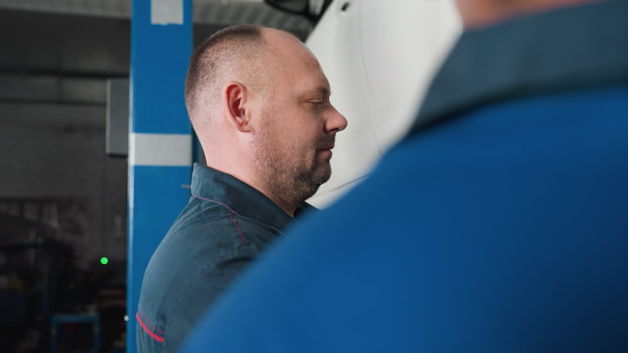 vista trasera de un mecánico en uniforme azul viendo a su colega usando una herramienta neumática para apretar el perno de un coche en un garaje, el mecánico mira hacia atrás mientras observa el proceso de reparación de la rueda