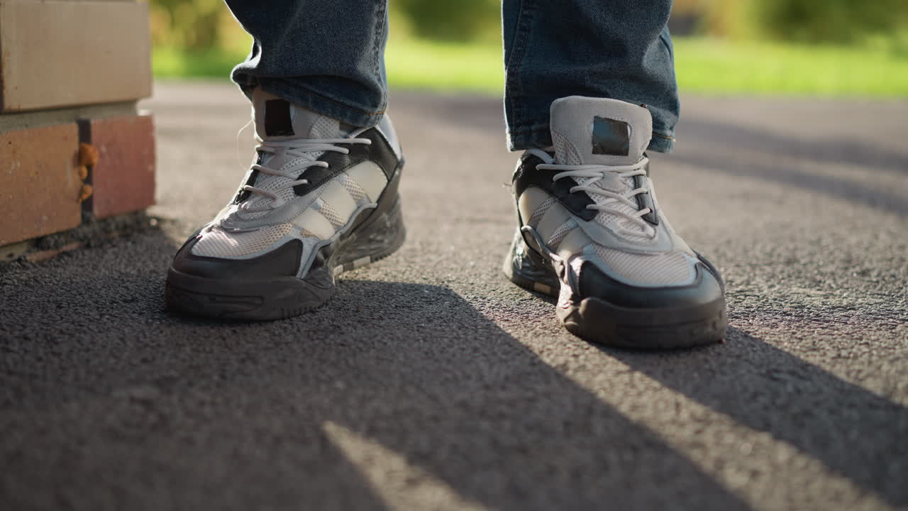 leg view of man in jeans and sneakers gently tapping right foot on asphalt with strong sunlight casting long shadow near brick wall and blurred greenery in background