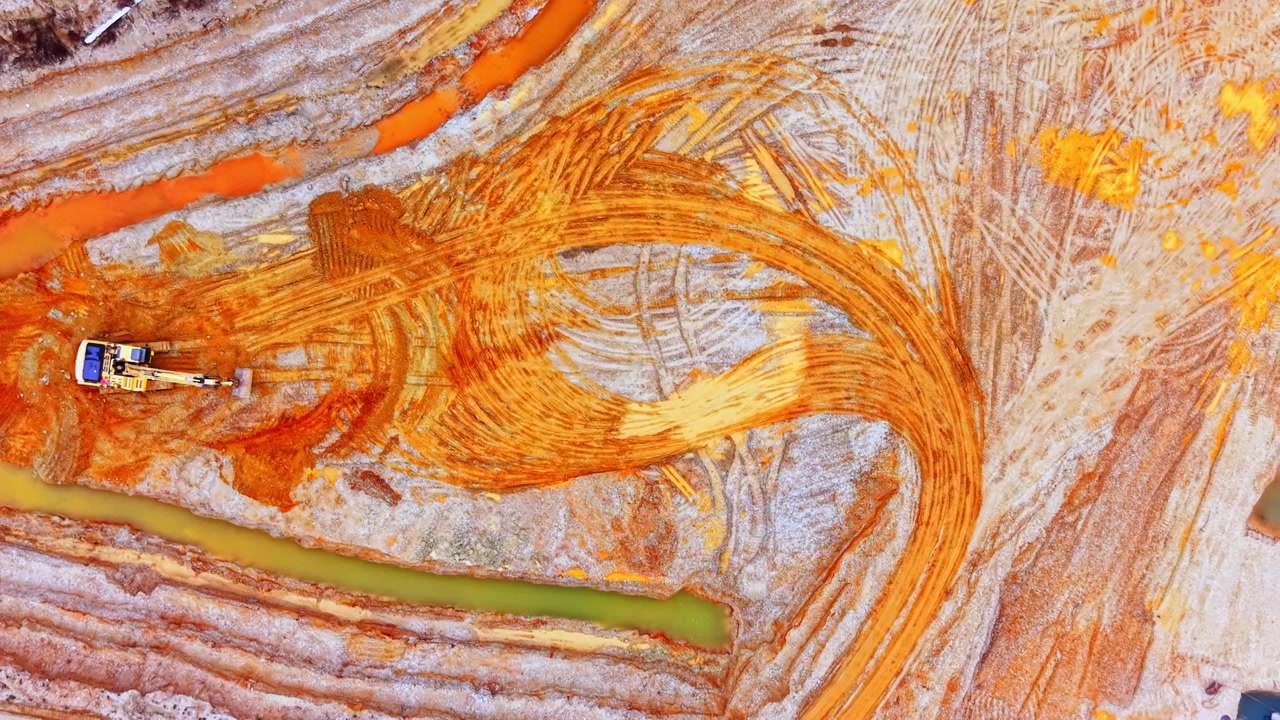 Bagger bei der Arbeit in farbenfrohen Sandgrubenmustern. Luftaufnahme zeigt wirbelnde orange und gelbe Spuren, die in nassen Mineralsand gegraben sind.