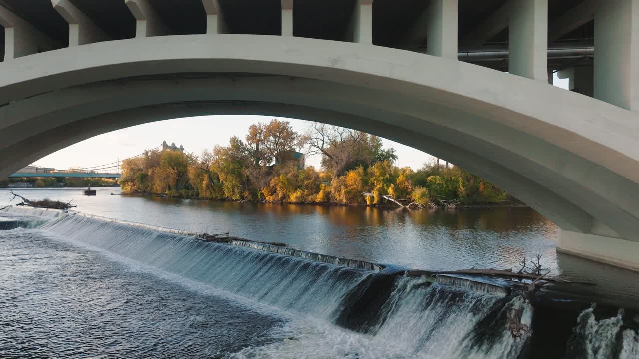 Low aerial shot flying under the Third Avenue Bridge arch, focusing on the St. Anthony Falls Dam spillway and fall foliage on the Mississippi River