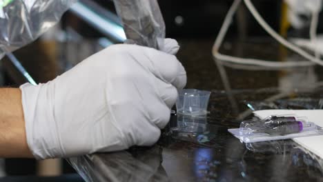 Tattoo-cups-with-black-ink-in-them-as-artist-plastic-gloved-hand-dips-needle-tip-and-sucks-up-liquid,-with-two-unopened-needle-tips-in-package-and-cling-wrap-desk