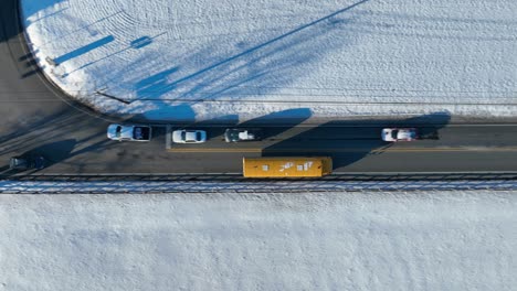 Top-down-aerial-tracking-shot-of-yellow-school-bus-driving-on-rural-road-surrounded-by-snow-covered-landscape-in-winter