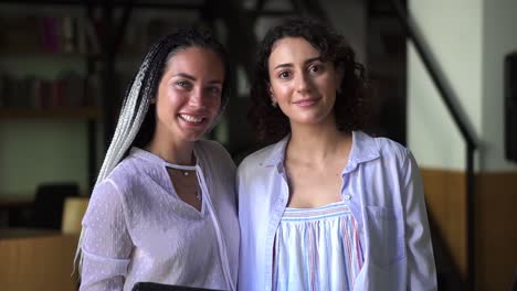 Portrait-of-two-pretty-young-caucasian-women-in-white-blouses-standing-in-library-and-smiling-in-camera,-academic-friends