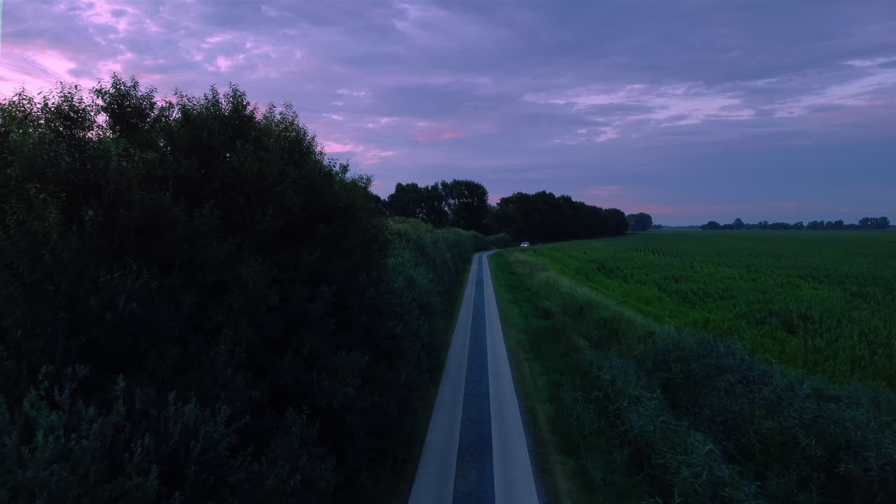 un coche conduciendo en una carretera entre un prado y un campo de maíz, disparo de drone de mal humor