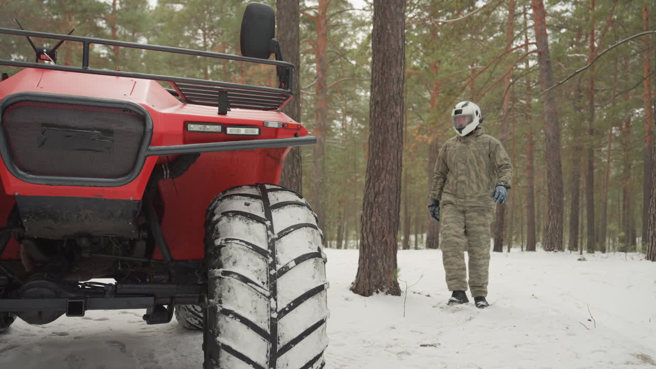 Un pilota si avvicina a un ATV rosso nella neve, appoggia la mano guantata sulla ruota anteriore, ispeziona il battistrada e la griglia, scruta la pineta circostante, in uno stato d'animo calmo e cauto, preparandosi per un percorso fuoristrada estremo.