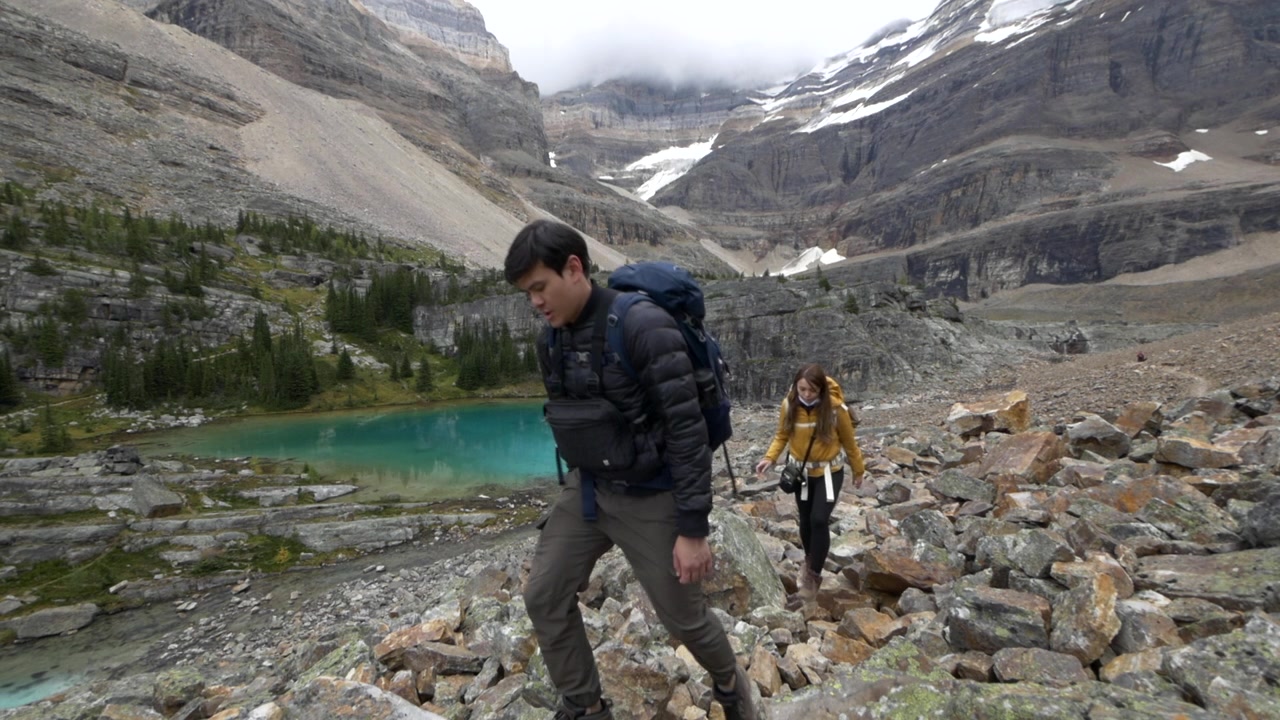 Una giovane coppia asiatica che sale attraverso un campo di massi sopra le rive turchesi del Lago O'Hara, Alberta, Canada