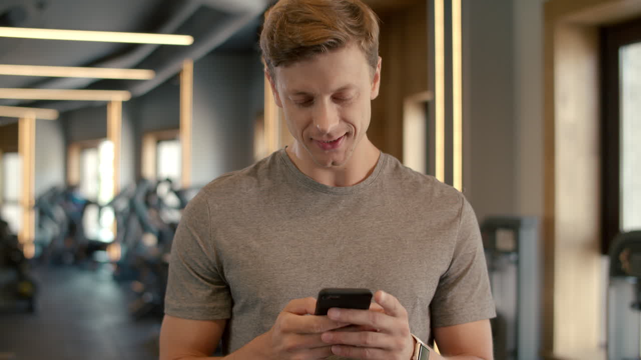 Cheerful bodybuilder standing at gym. Sportsman texting message in sport club