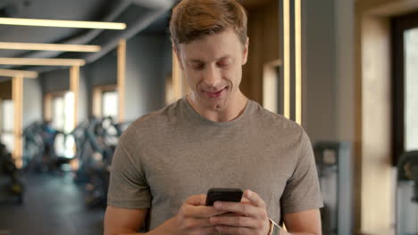 Cheerful-bodybuilder-standing-at-gym.-Sportsman-texting-message-in-sport-club